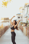 Model wearing Aura7 Activewear crisscross Malibu sports bra smiling with arms up on a sidewalk near the beach