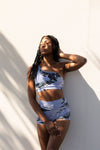Model wearing Aura 7 Activewear Tropic Rigel Short posing outdoors in the shade against a white wall background