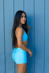 model wearing Aura 7 Activewear Fresh Air Rigel Short posing outdoors against a blue panel wall
